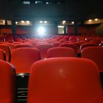 Sala de cinema vazia, com fileiras de poltronas vermelhas voltadas para uma tela iluminada
