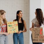 Três mulheres seguram cartazes de apoio aos direitos das mulheres em um ambiente interno.