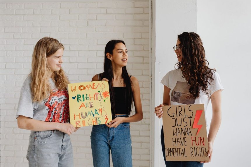 Três mulheres seguram cartazes de apoio aos direitos das mulheres em um ambiente interno.