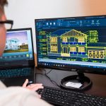 Arquiteto analisa projetos detalhados de uma residência exibidos em dois monitores, em um escritório moderno.