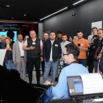Público diverso acompanha atentamente uma demonstração de piano em um ambiente interno.