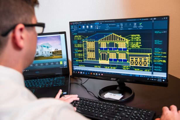 Arquiteto analisa projetos detalhados de uma residência exibidos em dois monitores, em um escritório moderno.